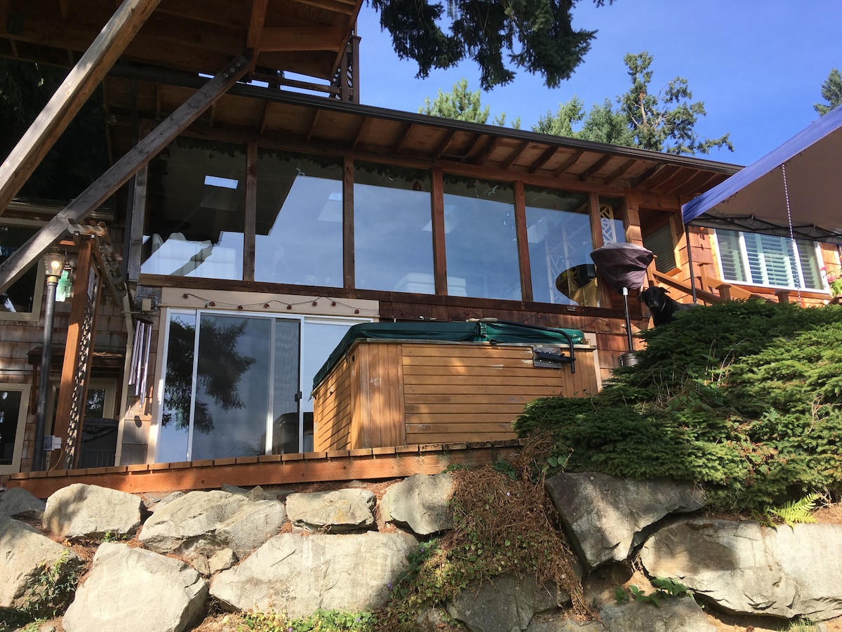 A wooden structure is shown, highlighted by large glass windows that provide views of the surroundings. A hot tub is positioned on a wooden deck, surrounded by natural stone landscaping. The exterior showcases rich wood tones against green foliage, merging with the natural environment.