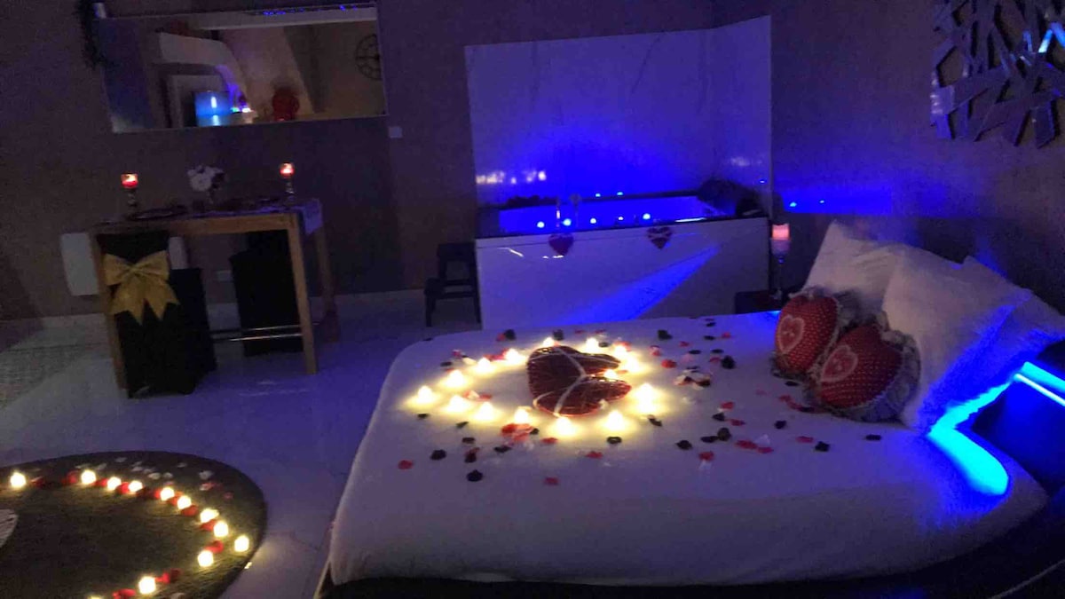 A spacious bed is adorned with red heart-shaped cushions and surrounded by soft, glowing lights. A romantic arrangement of rose petals and candles enhances the atmosphere. In the background, a modern jacuzzi is visible, highlighted by subtle blue and white lighting.