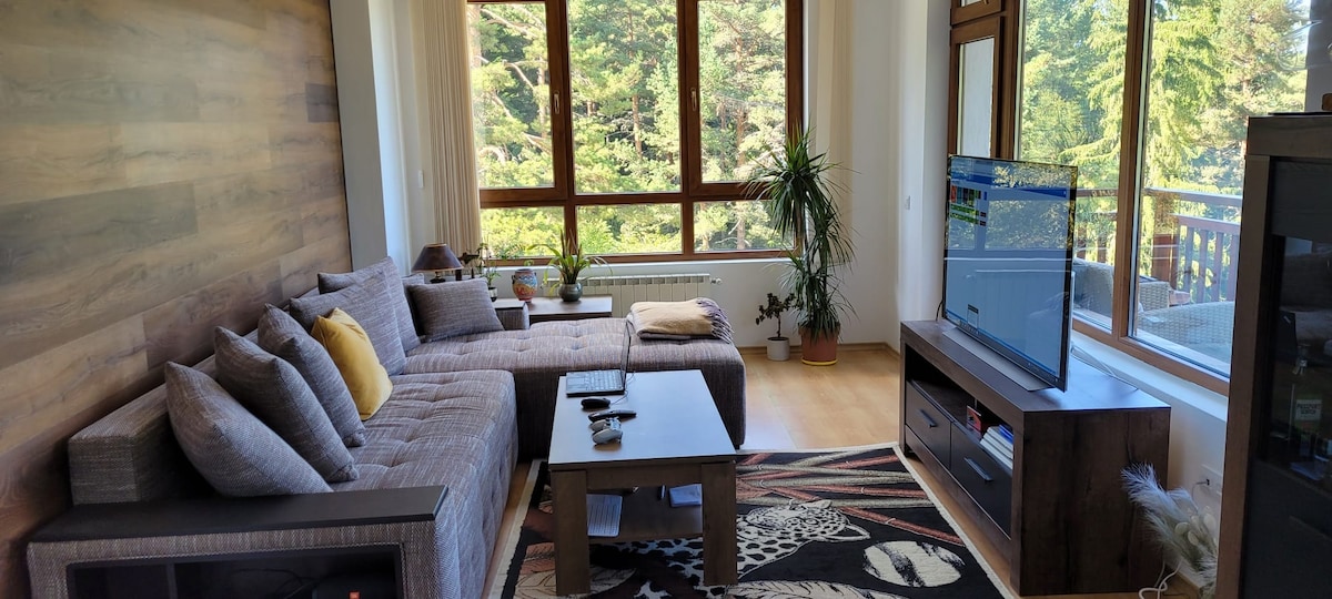 A spacious living area features a large sectional sofa adorned with pillows and a coffee table. Natural light fills the space through a series of large windows, offering views of surrounding greenery. A sleek television is mounted on a dark wooden media unit, enhancing the modern aesthetic.