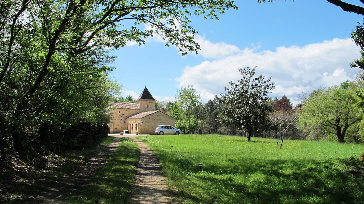 A scenic view along a pathway leads to a charming stone house surrounded by lush greenery. The sky is bright with clouds, and a car is parked near the entrance. Trees frame the area, enhancing the peaceful rural setting.