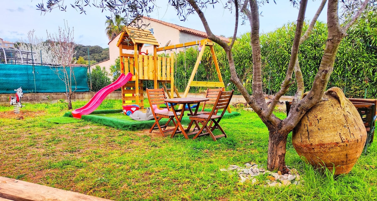 A spacious outdoor area features a children’s playset with a slide and swing, alongside a wooden dining table and chairs. Lush green grass is surrounded by shrubs and trees, providing a serene environment. A large decorative pot is positioned nearby.