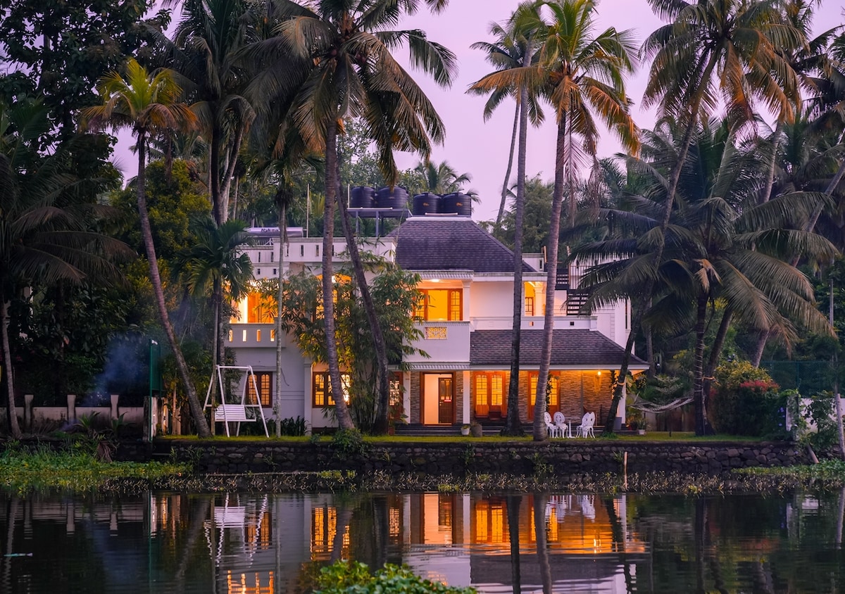A charming three-story house is nestled among palm trees, facing a serene lake. Warm lights glow from large windows, reflecting off the water. A small porch with white chairs is visible in the foreground, enhancing the tranquil lakeside setting.