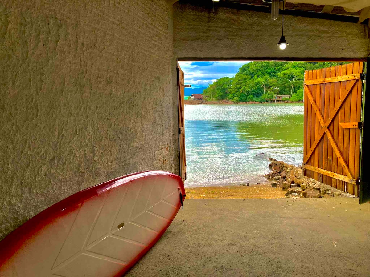 An entryway leading to a serene body of water is visible, framed by rustic wooden doors. A stand-up paddleboard rests against the wall, and the gentle ripples of the water reflect the greenery in the distance.