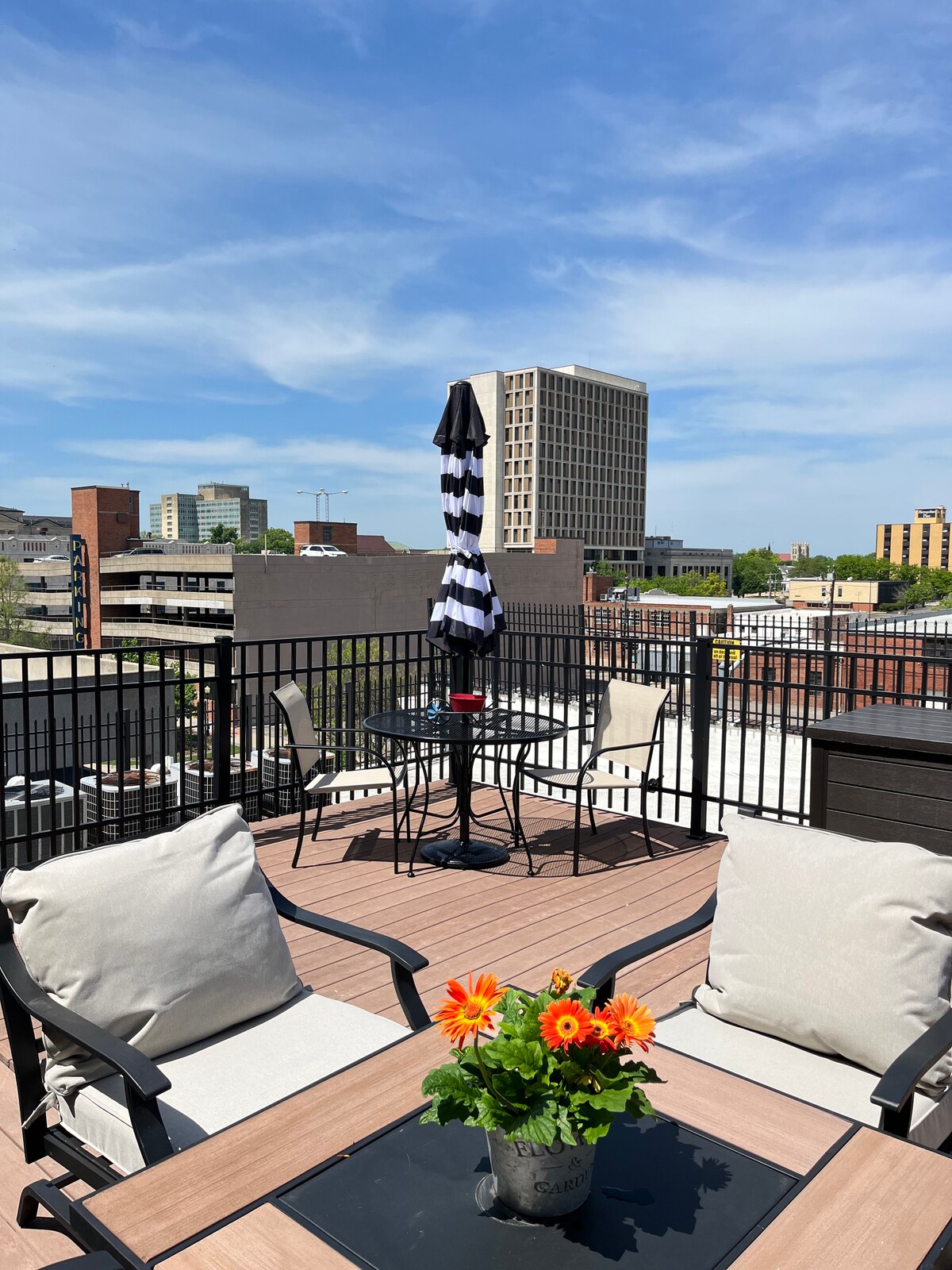 A rooftop deck offers sweeping views of Downtown Topeka, featuring a black and white striped umbrella over a dining set. Two comfortable chairs frame a table with a flower arrangement, inviting relaxation under a clear blue sky.