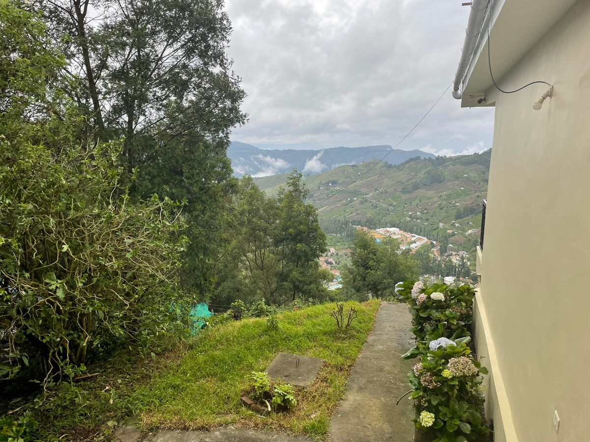 A scenic view captures the rolling hills and valley beyond, with greenery lining the foreground. The path is bordered by shrubs and flowers, leading to a tranquil outdoor space. Overcast skies create a serene atmosphere, enhancing the natural beauty of the surroundings.