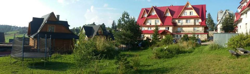 A panoramic view of the property displays a mix of buildings with vibrant red roofs and natural greenery. Adjacent to the main structure, a trampoline is visible, surrounded by lush grass and trees. The landscape feels spacious, offering a relaxing outdoor environment.