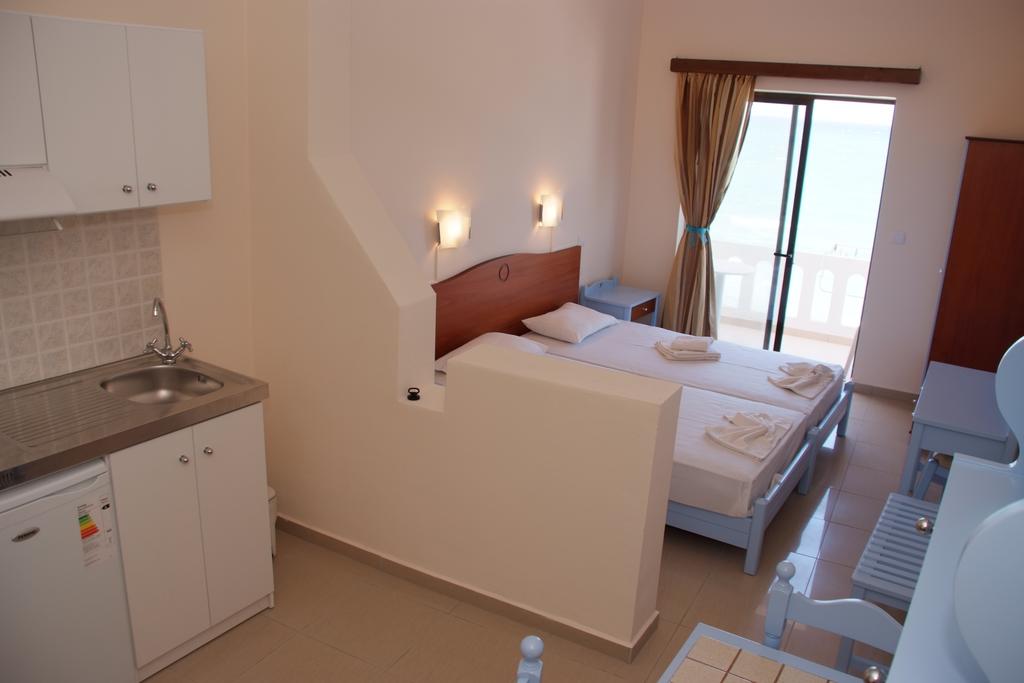 The studio features two single beds separated by a partition, with light streaming in through a sliding glass door. A compact kitchenette is situated in one corner, and a wardrobe is visible along the wall. Soft colors are present throughout the space, enhancing the coastal ambiance.