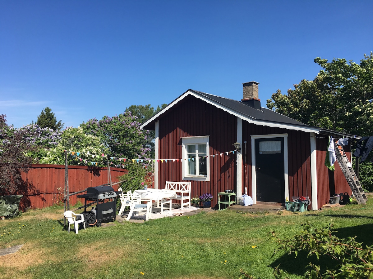 A charming red cottage with a black roof is surrounded by a well-maintained garden. A patio area features white outdoor furniture, while a barbecue grill is visible nearby. Lush greenery and blooming flowers add to the inviting atmosphere of the outdoor space.