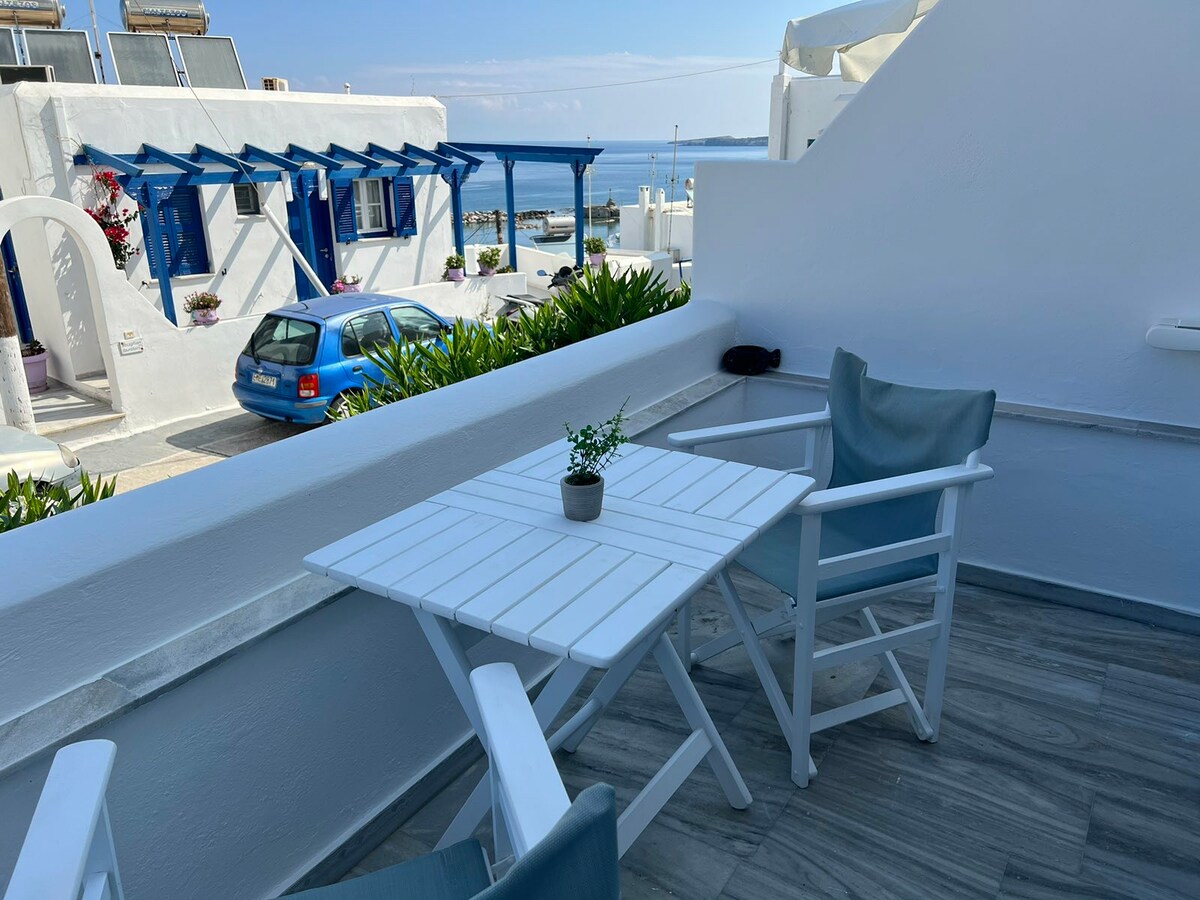A private balcony features a white wooden table and two light blue chairs. Potted greenery sits at the center of the table. In the background, a glimpse of the sea and a blue car is visible, framed by the whitewashed architecture.