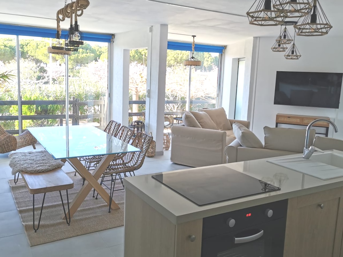 A spacious living area is highlighted by large windows, allowing natural light to fill the space. A glass dining table is paired with woven chairs. The open kitchen features modern appliances and an island with a stovetop, while comfortable seating is positioned nearby.