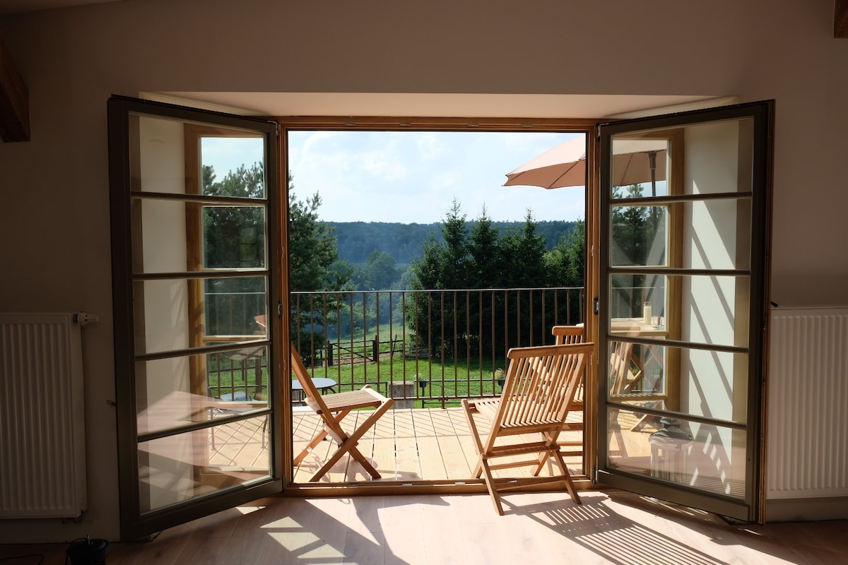Double French doors open to a spacious balcony overlooking lush green trees and rolling hills. Two wooden chairs are positioned on the balcony, inviting guests to relax and enjoy the scenic view. Natural light filters through the doors, highlighting the interior space.