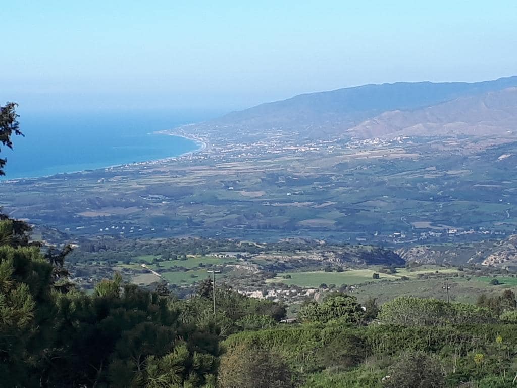 A sweeping coastal view is captured, showcasing the turquoise waters of the sea meeting the shoreline. Rolling hills and lush fields create a vibrant landscape, complemented by distant mountains under a clear blue sky.
