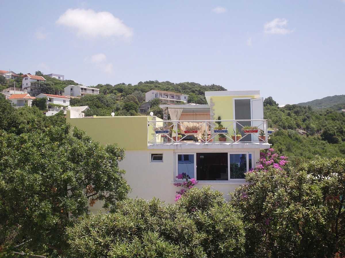 A three-story villa with bright yellow accents is embraced by lush greenery. The outdoor terrace is adorned with flowering plants, offering an inviting space for relaxation. The surrounding hillside provides a scenic backdrop, enhancing the overall tranquil setting.