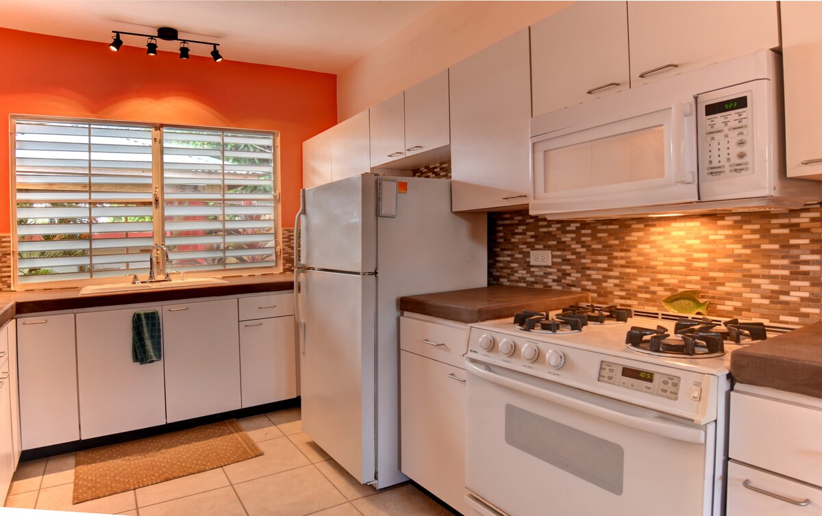 A bright kitchen features white cabinetry and modern appliances, including a gas stove and refrigerator. Natural light enters through a window with slatted shutters, showcasing a stylish backsplash. The space is complemented by a small rug in front of the sink area.