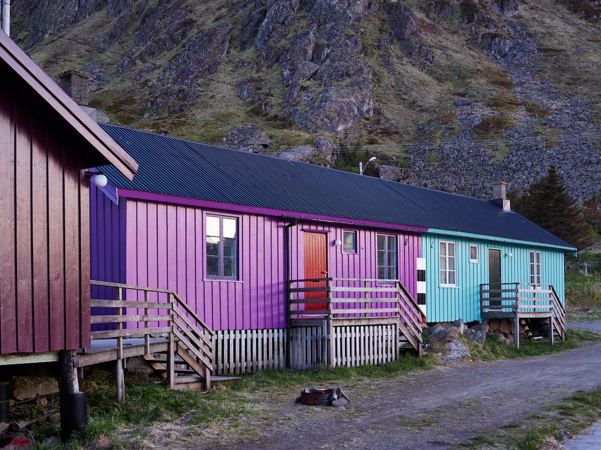 A row of rustic cabins with colorful exteriors in shades of pink, purple, and teal is presented. Each cabin features a wooden deck with steps leading up. A dirt path runs beside the cabins, bordered by grass and rocky terrain.