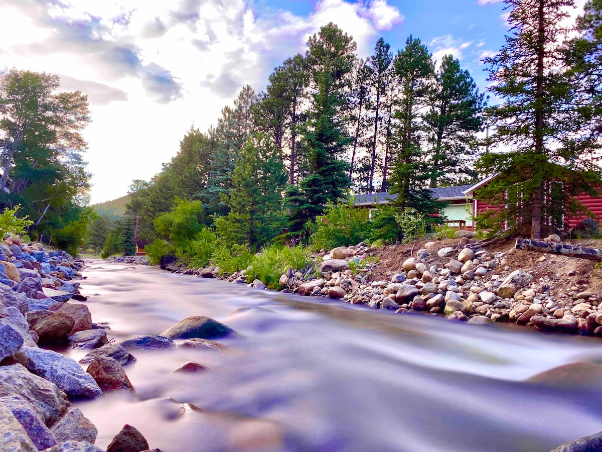 A tranquil river flows gently past smooth stones and vibrant greenery. Tall pine trees line the riverbank, framing a charming red house nestled among the foliage. The scene captures a peaceful natural setting, inviting relaxation and reflection in a serene environment.
