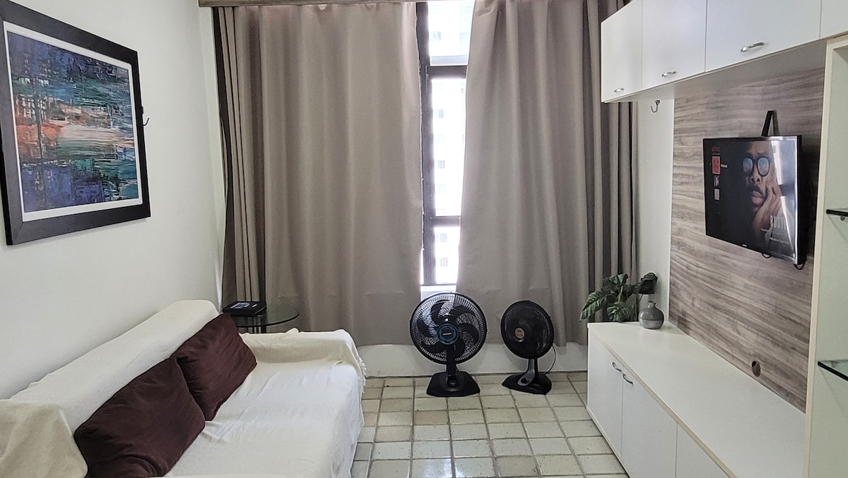 A living area features a comfortable white sofa with brown cushions and a mounted television. Two table fans are positioned nearby, and natural light filters through tall windows draped with light curtains, enhancing the bright and airy ambiance.