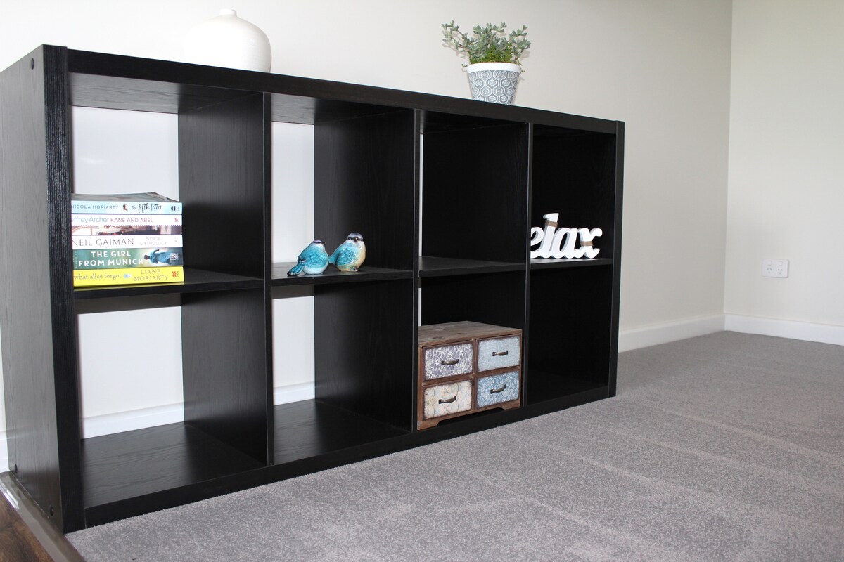 A modern black bookshelf is positioned against a light-colored wall, featuring several open compartments. Decorative items, including colorful books, a small plant in a pot, and two bird figurines, are displayed on top. A box with drawers adds an additional decorative touch.