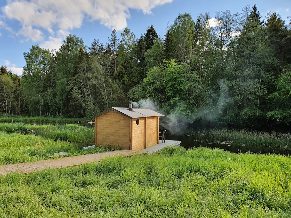 A wooden sauna is positioned near a river, surrounded by lush greenery. Smoke gently rises from the chimney, blending with the serene landscape. A wooden path leads from the sauna to the grassy riverbank, inviting a calm retreat in nature.