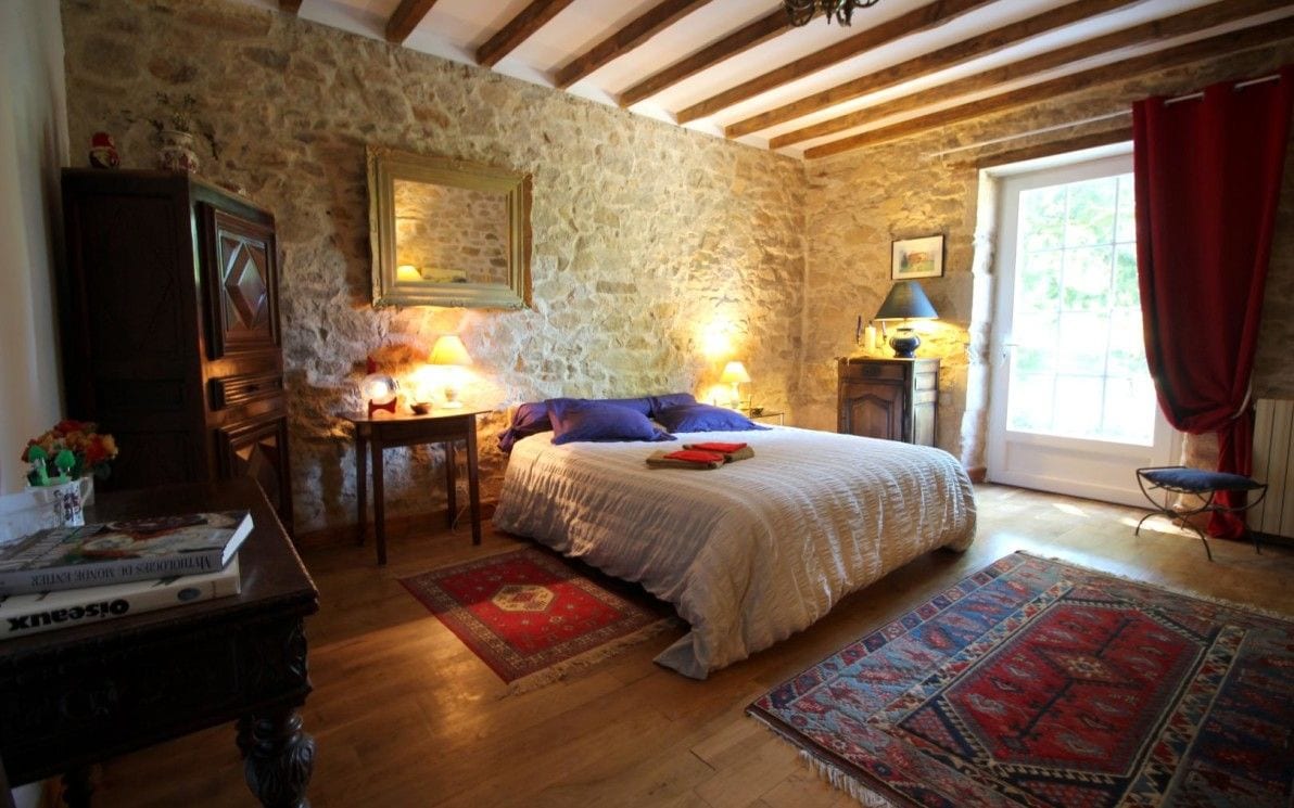 A spacious bedroom features stone walls and wooden beams overhead. A large, well-made bed is positioned centrally, flanked by bedside tables with lamps. A cozy rug adds warmth to the wooden floor, while a large window lets in natural light, framed by red curtains.