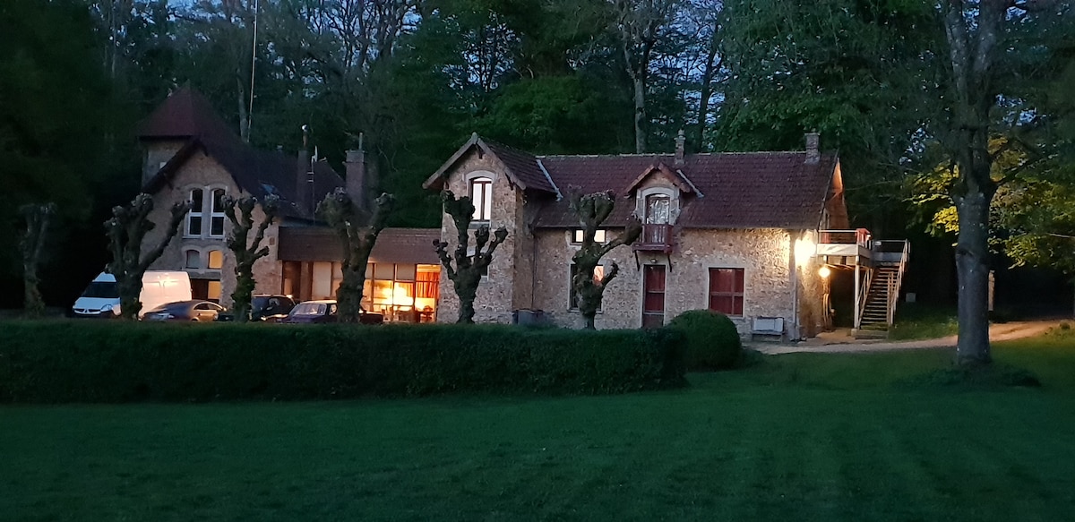 A charming stone structure is illuminated softly in the evening light, blending with the surrounding greenery. Trees are neatly trimmed, and a pathway leads to the entrance. The building's rustic architecture features multiple windows and a balcony area overlooking the landscaped lawn.