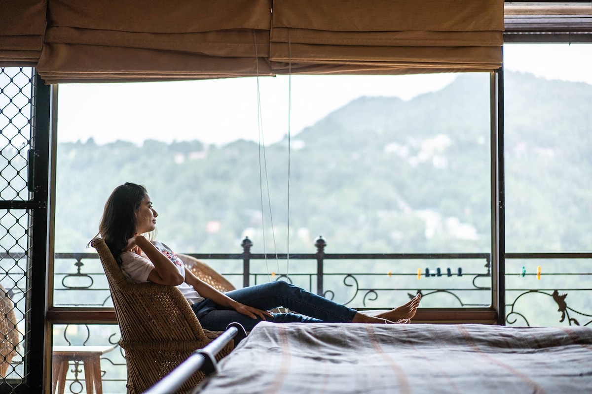 A comfortable seating area is set beside large glass windows, revealing expansive views of the surrounding hills. A figure relaxes in a woven chair, while soft natural light filters through the window coverings, creating a serene ambiance in the space.