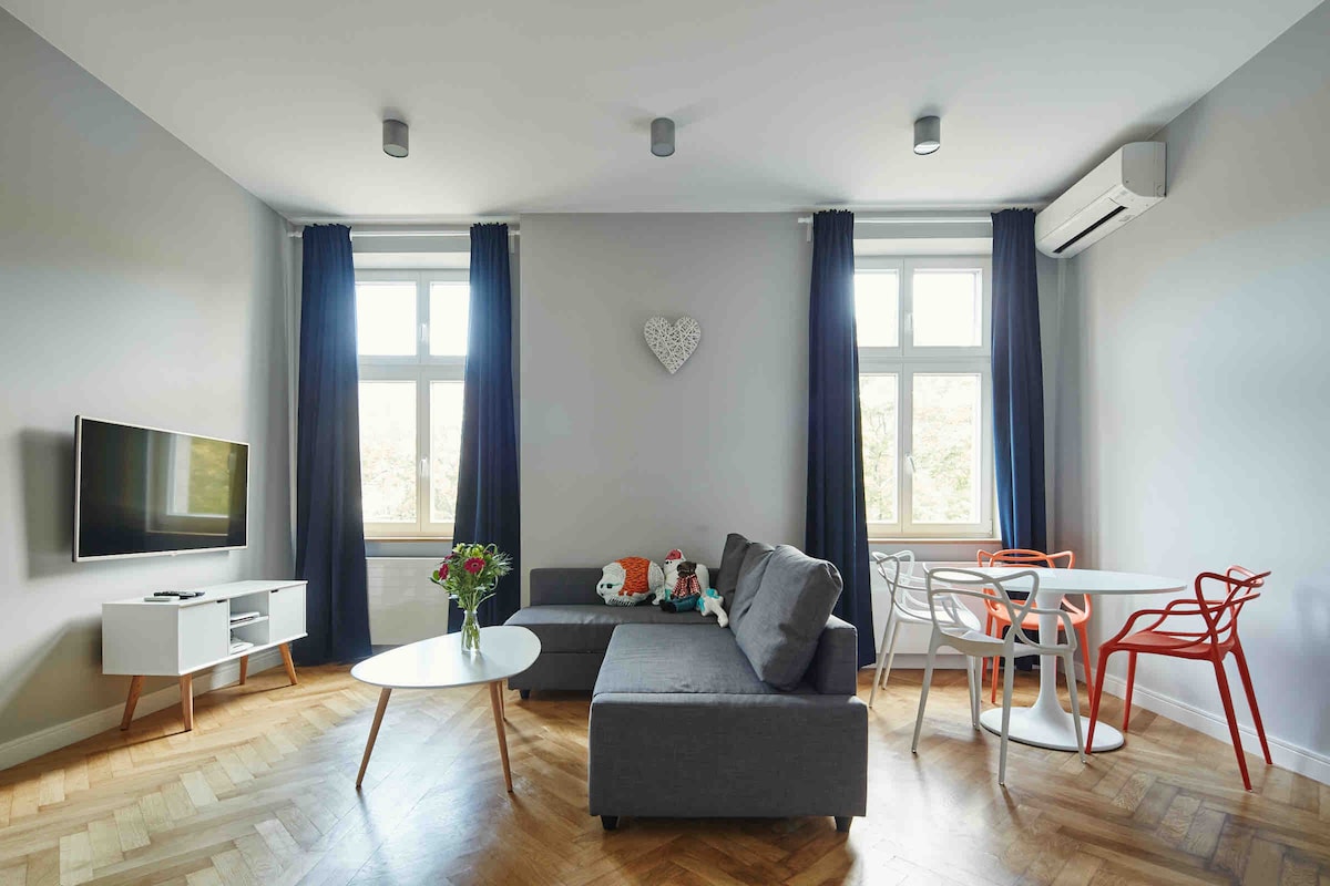 A modern living area is featured, showcasing a spacious gray sofa set against light walls. Natural light enters through two large windows, paired with dark curtains. A circular white table is surrounded by colorful chairs, while a television and decorative elements add to the vibrant ambiance.
