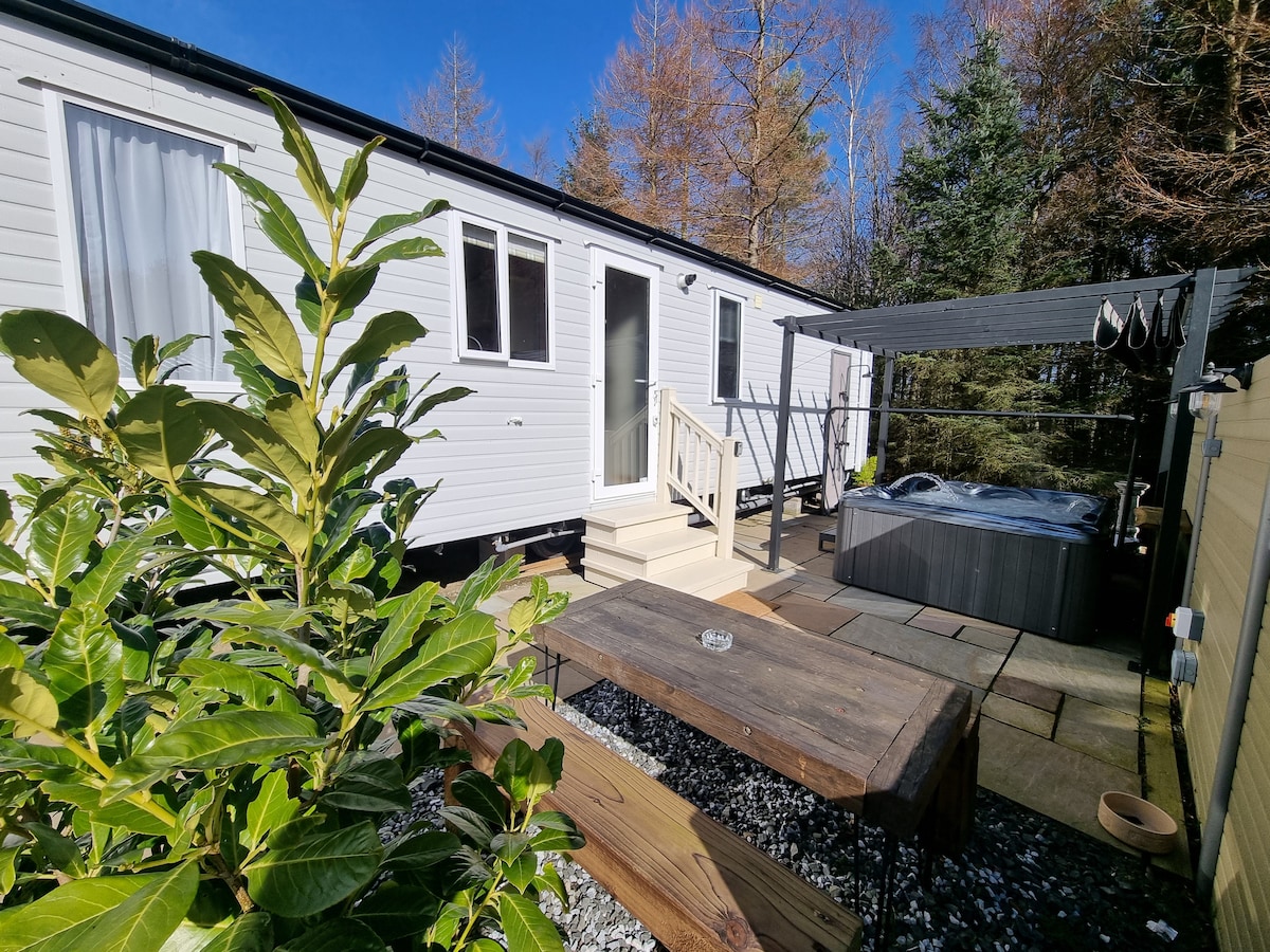A lodge exterior is shown, featuring light-colored siding and a welcoming entrance. A private hot tub is positioned under a covered area, and a wooden table is set on a stone patio. Lush greenery surrounds the space, complementing the natural setting.
