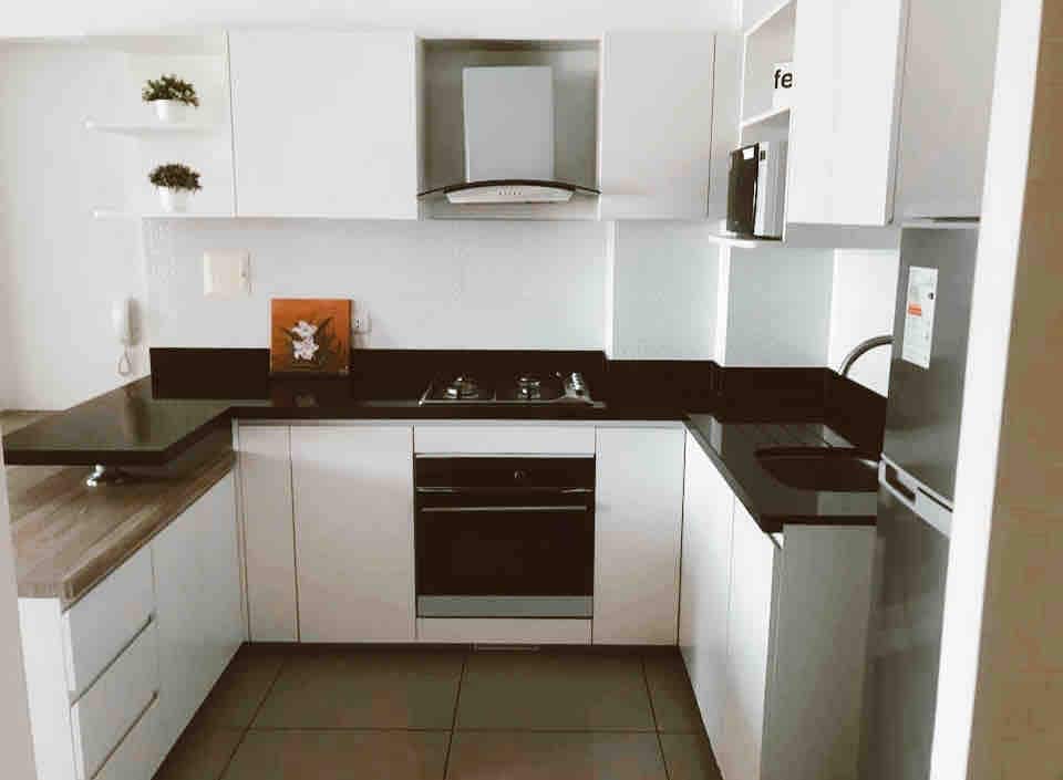 A modern kitchen features sleek white cabinets contrasted by dark countertops. A stove and oven are integrated into the cabinetry, with an extractor hood positioned above. A refrigerator stands in the corner, and open shelves display small plants and decorative items.