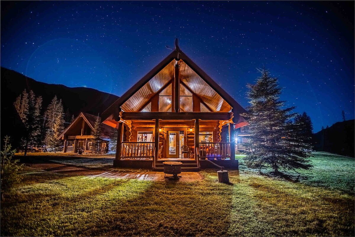 An inviting log cabin is highlighted by warm outdoor lighting beneath a clear night sky filled with stars. The spacious covered porch displays wooden railings and is surrounded by well-kept grass and tall trees, creating a peaceful retreat in nature.