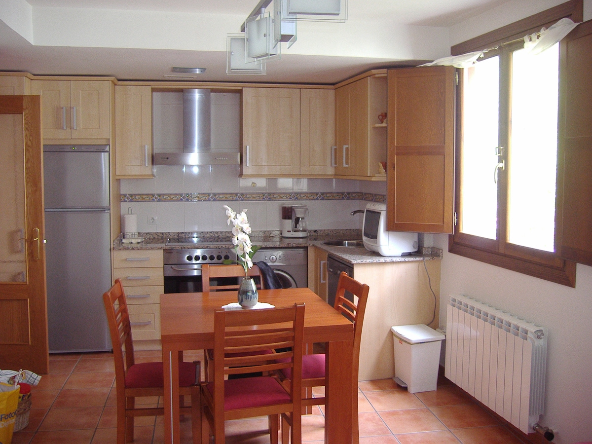 The kitchen area features light wood cabinetry and a granite countertop. A dining table with four wooden chairs is positioned in the center. Modern appliances include a refrigerator, oven, and microwave. Natural light enters through a window above the sink, providing an airy feel to the space.