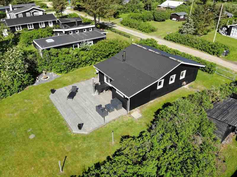 A modern summerhouse is surrounded by greenery, featuring a spacious wooden terrace with outdoor seating. The well-maintained lawn offers ample space for relaxation. Nearby structures are visible, blending with the tranquil natural surroundings.