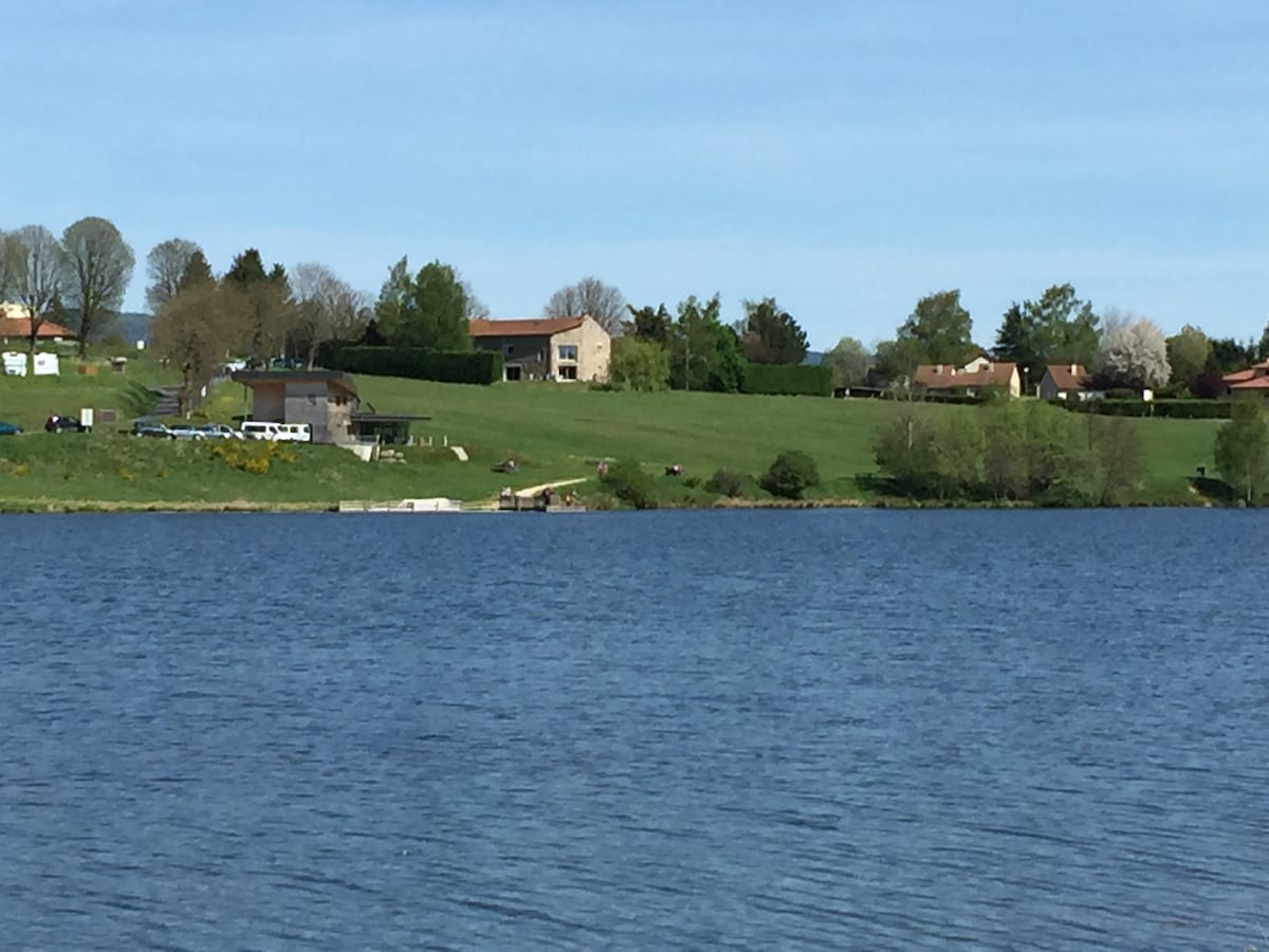 A serene landscape features a calm lake surrounded by lush greenery and gently rolling hills. Several buildings, including residential homes and a smaller facility, are visible on the opposite shore, with trees and flowering plants providing a natural backdrop.