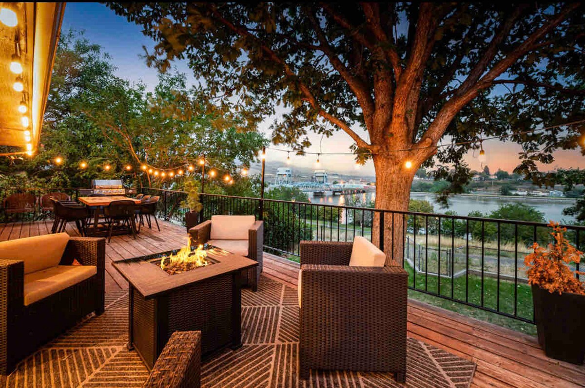 A private deck is featured, adorned with string lights and surrounded by trees. Cozy seating areas with cushioned chairs and a central fire pit provide a welcoming space for relaxation. Unobstructed views of the river can be enjoyed from the deck, enhancing outdoor experiences.