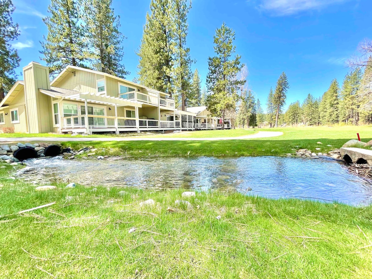 A two-story townhome is situated next to a gently flowing stream on a golf course. The structure features a series of large windows, allowing natural light to fill the space. Lush green grass surrounds the home, framed by tall trees in the background.