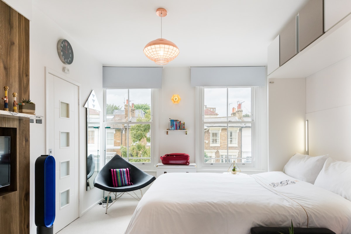 A bright studio features a spacious bed with crisp white linens, flanked by large windows. A modern light fixture hangs above, and decorative items are visible on shelves. A stylish black chair with colorful pillows offers additional seating, completing the inviting atmosphere.