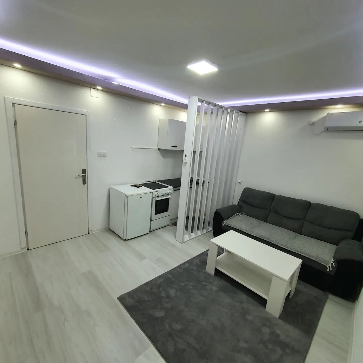 A modern living space features a cozy gray couch facing a light-colored coffee table. A compact kitchen area with essential appliances is visible behind a white partition. Soft lighting highlights the room's clean lines and neutral tones, creating a calm environment.