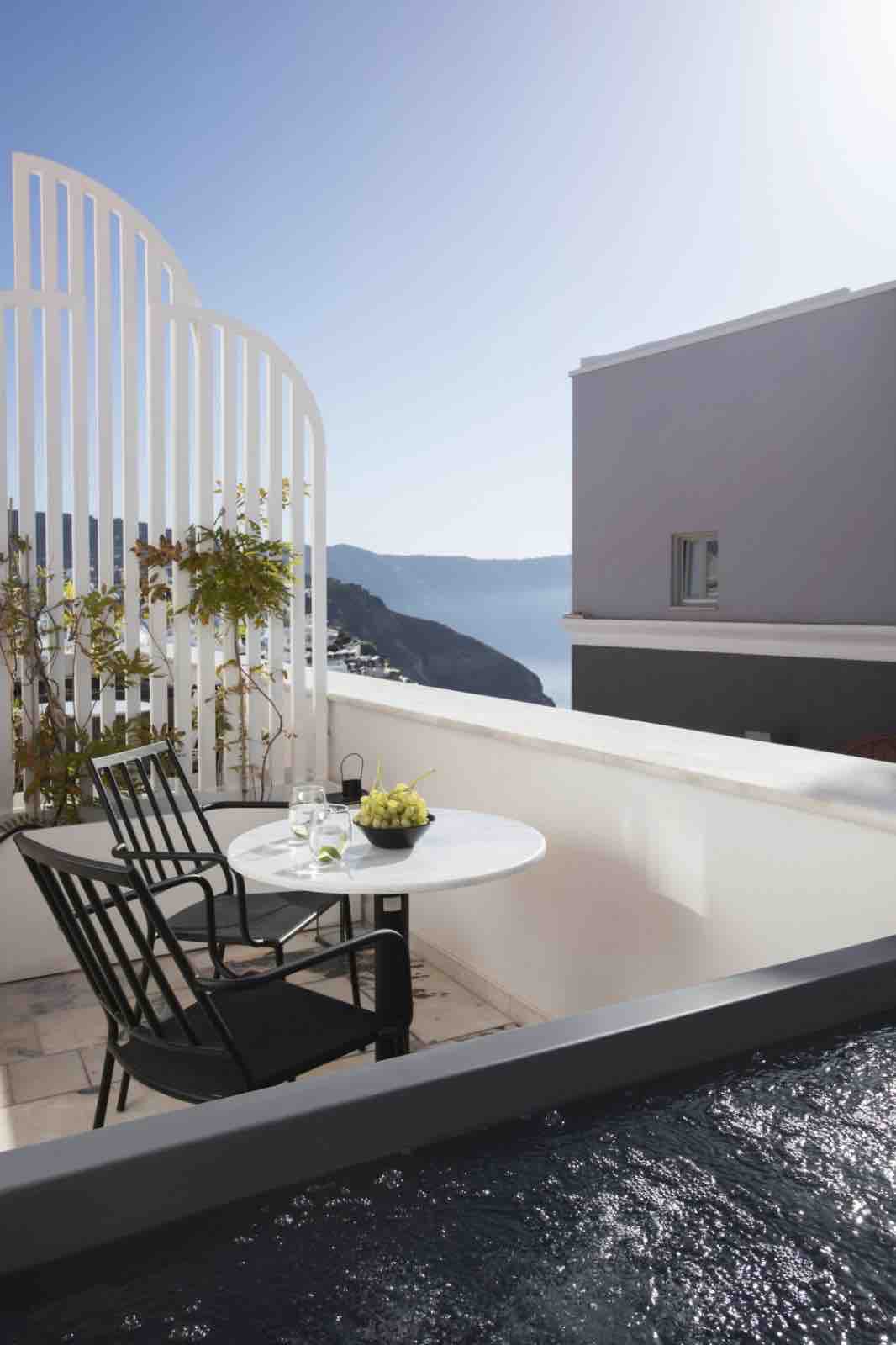 A small outdoor terrace is furnished with a round table and two black chairs. Potted plants are growing on the white trellis, and the ocean view is visible in the background, complemented by the bright blue sky.