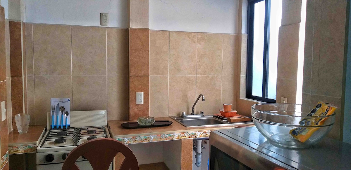 A functional kitchen area features a gas stove, a large sink, and ample counter space. Natural light enters through a window, highlighting kitchenware arranged on the countertop. A dining chair is positioned nearby, adding to the inviting atmosphere of the space.