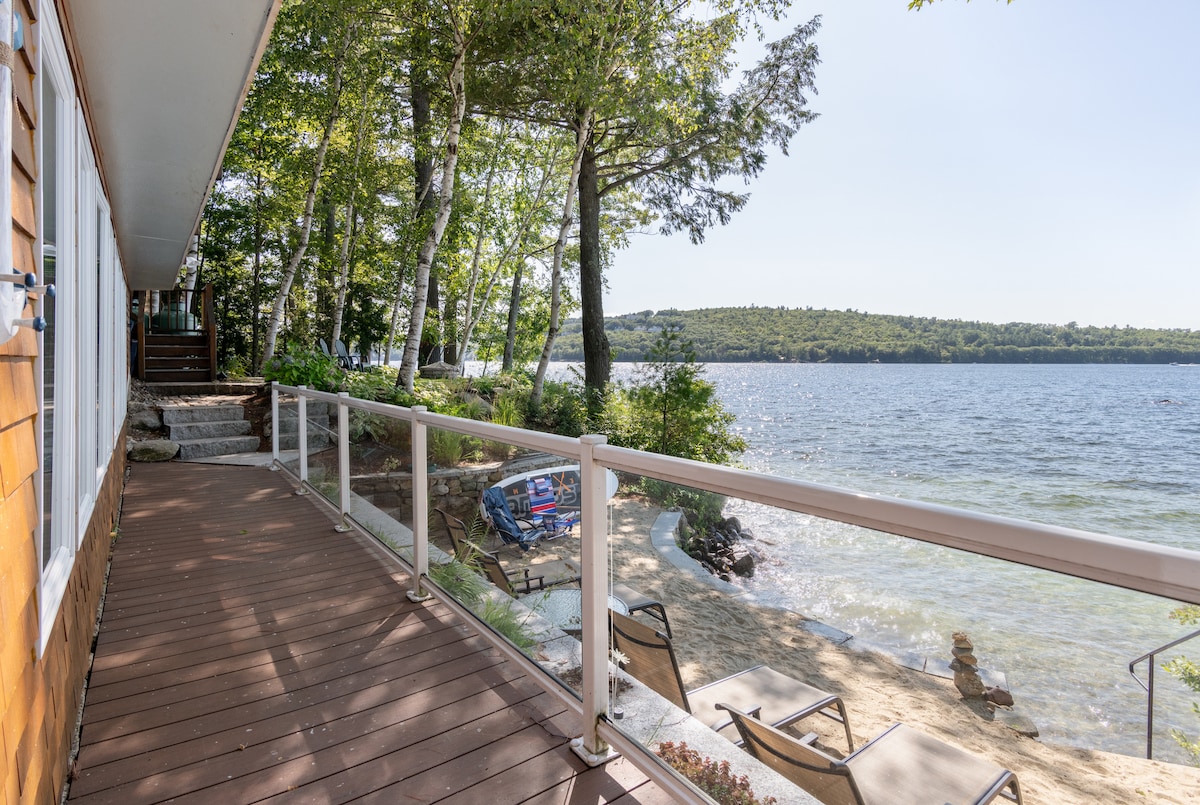 A wooden deck extends along the waterfront, offering views of the tranquil lake and surrounding greenery. Lounge chairs are positioned at the edge, inviting relaxation. Sunlight reflects on the water, enhancing the serene outdoor space.