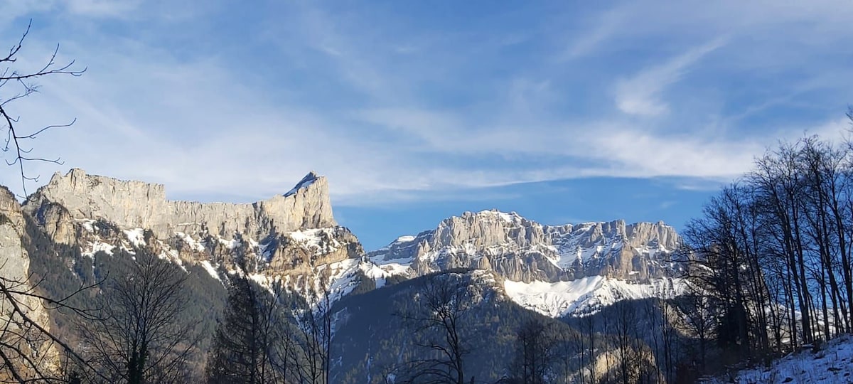 A panoramic view of mountain ranges under a clear blue sky is presented. Snow-capped peaks and rugged cliffs are visible, surrounded by trees with bare branches. The serene landscape reflects a natural setting, inviting exploration and appreciation of the surroundings.