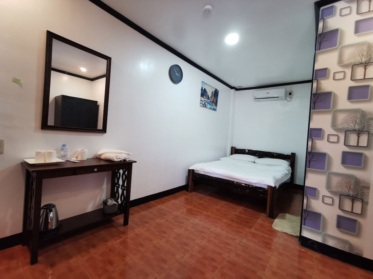 An inviting room features a spacious bed with white linens, a wall-mounted air conditioner, and a modern cabinet. A dark wood table holds a kettle and towels, while a large mirror reflects the room's natural light. Decorative artwork adds subtle interest to the wall.