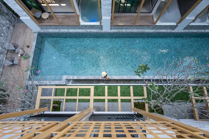 Aerial view of a serene swimming pool bordered by wooden decking and lush green landscaping. The clear water is visually complemented by the surrounding tropical plants, while a person relaxes peacefully nearby. A modern structure is visible, enhancing the overall tranquil setting.