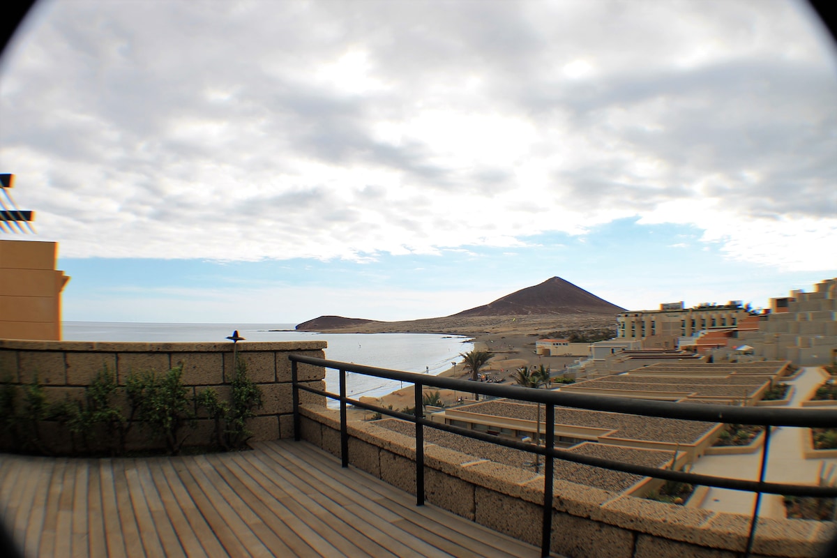A spacious terrace features a railing, providing unobstructed views of the coastline and Montaña Roja in the distance. The wooden deck is complemented by potted plants, enhancing the connection with the surrounding landscape. A cloudy sky creates a soft, muted backdrop.