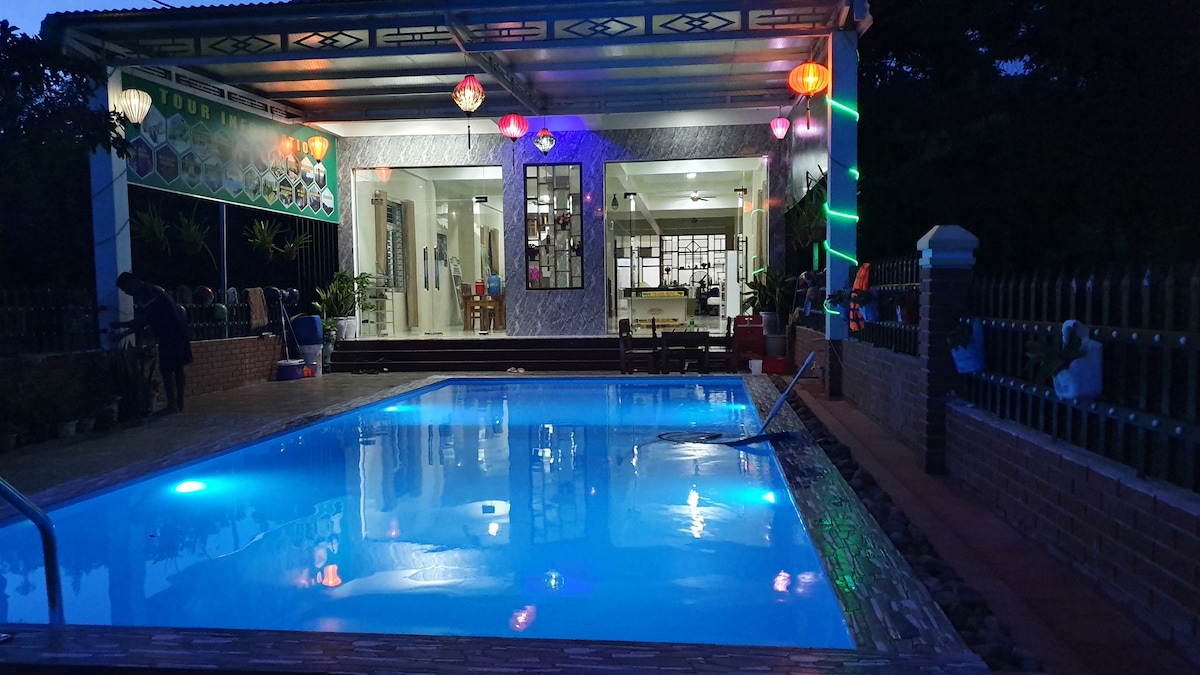 A well-lit outdoor area features a clear swimming pool surrounded by a paved deck. The main building showcases large windows and vibrant lanterns hanging from the ceiling. Lush greenery can be seen in the background, enhancing the serene atmosphere.