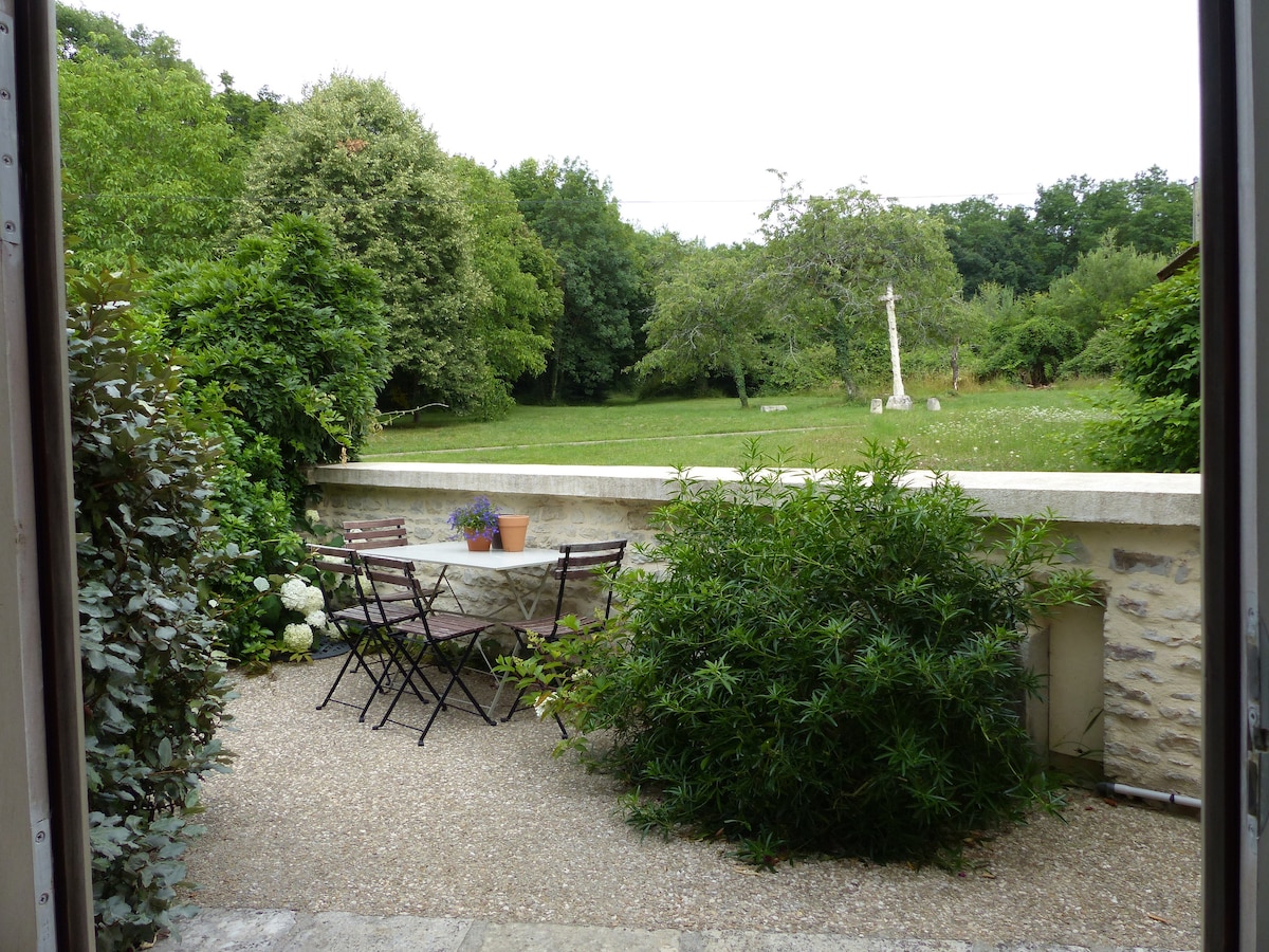 A peaceful outdoor terrace is shown, featuring a small table surrounded by folding chairs. Lush greenery and flowering plants create a natural border. Beyond the terrace, an expansive lawn with trees can be seen, extending into a wooded area.