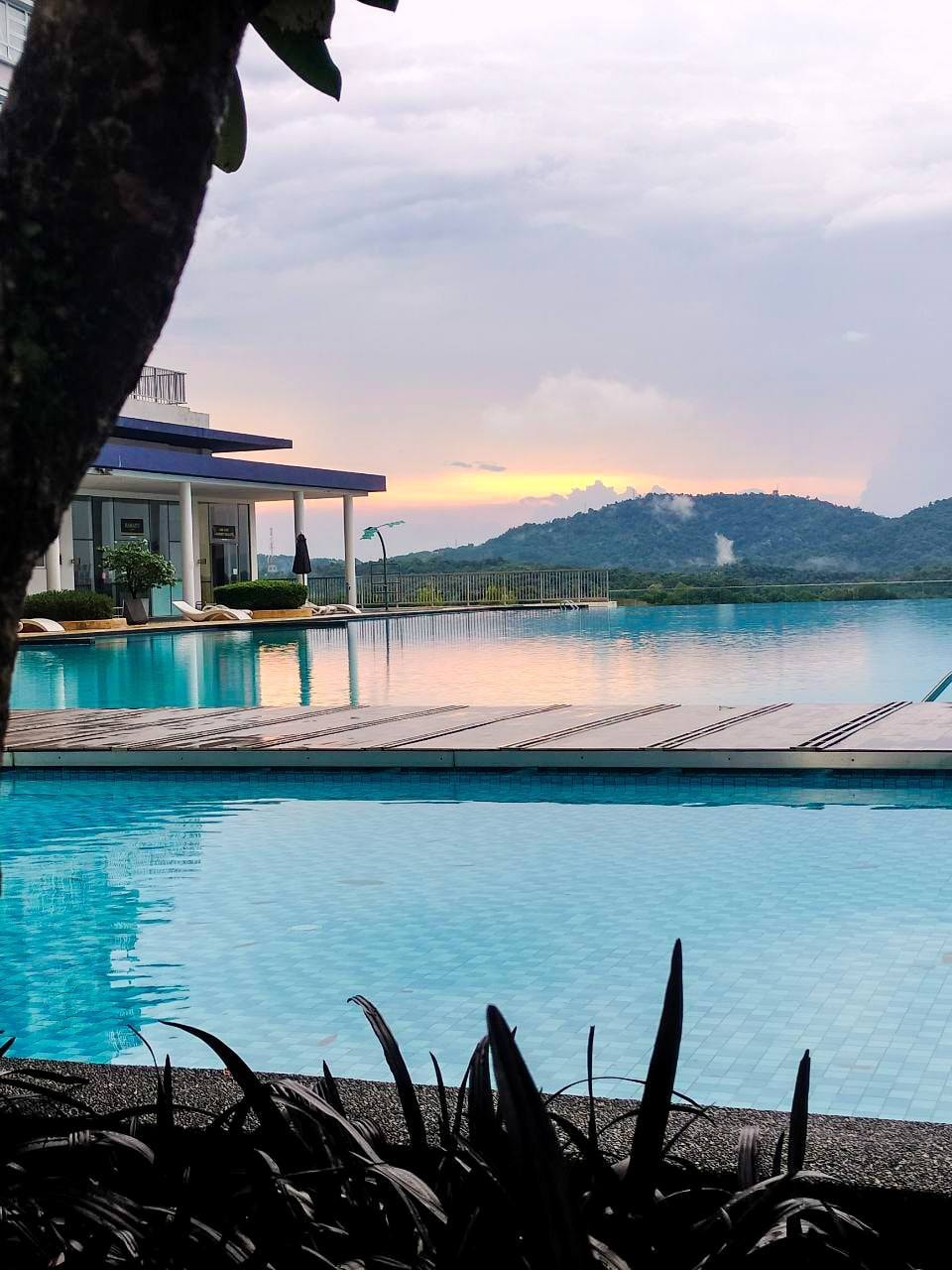 An infinity swimming pool is framed by lush green foliage and a wooden deck, with reflections of the surrounding landscape visible in the calm water. The distant mountains are partially shrouded in mist, adding a serene backdrop to the peaceful setting.