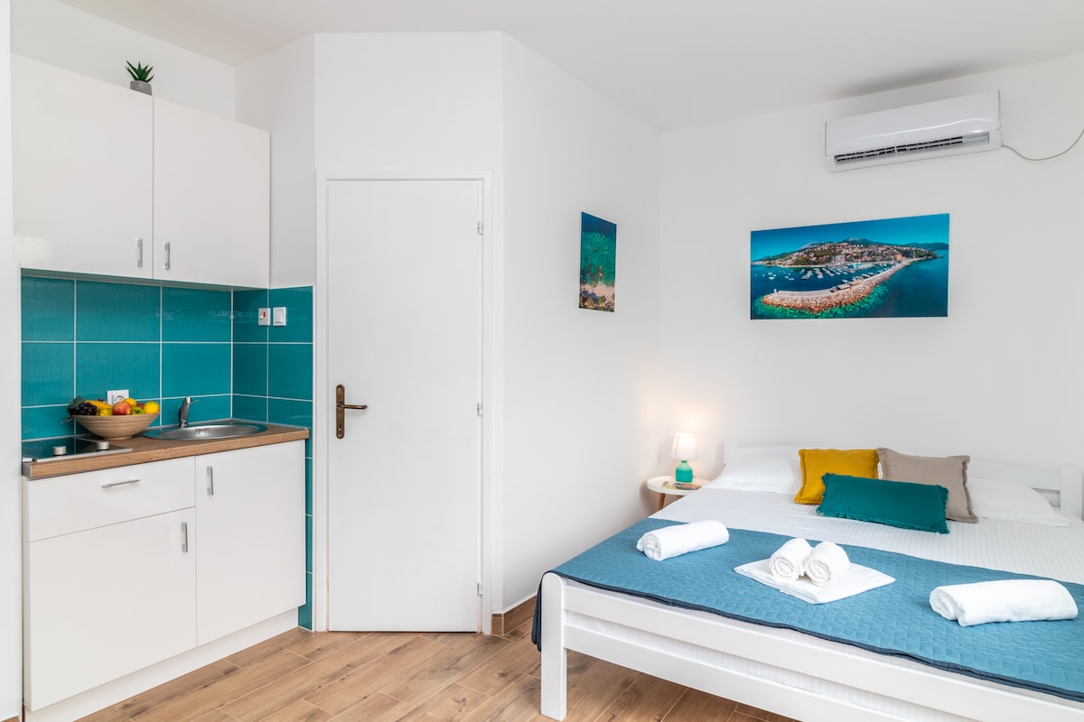A bright and airy room features a spacious bed with neatly arranged towels. A small kitchen area is visible, complete with a sink and countertop. Artwork adorns the walls, and natural light enhances the serene atmosphere, creating a welcoming space.