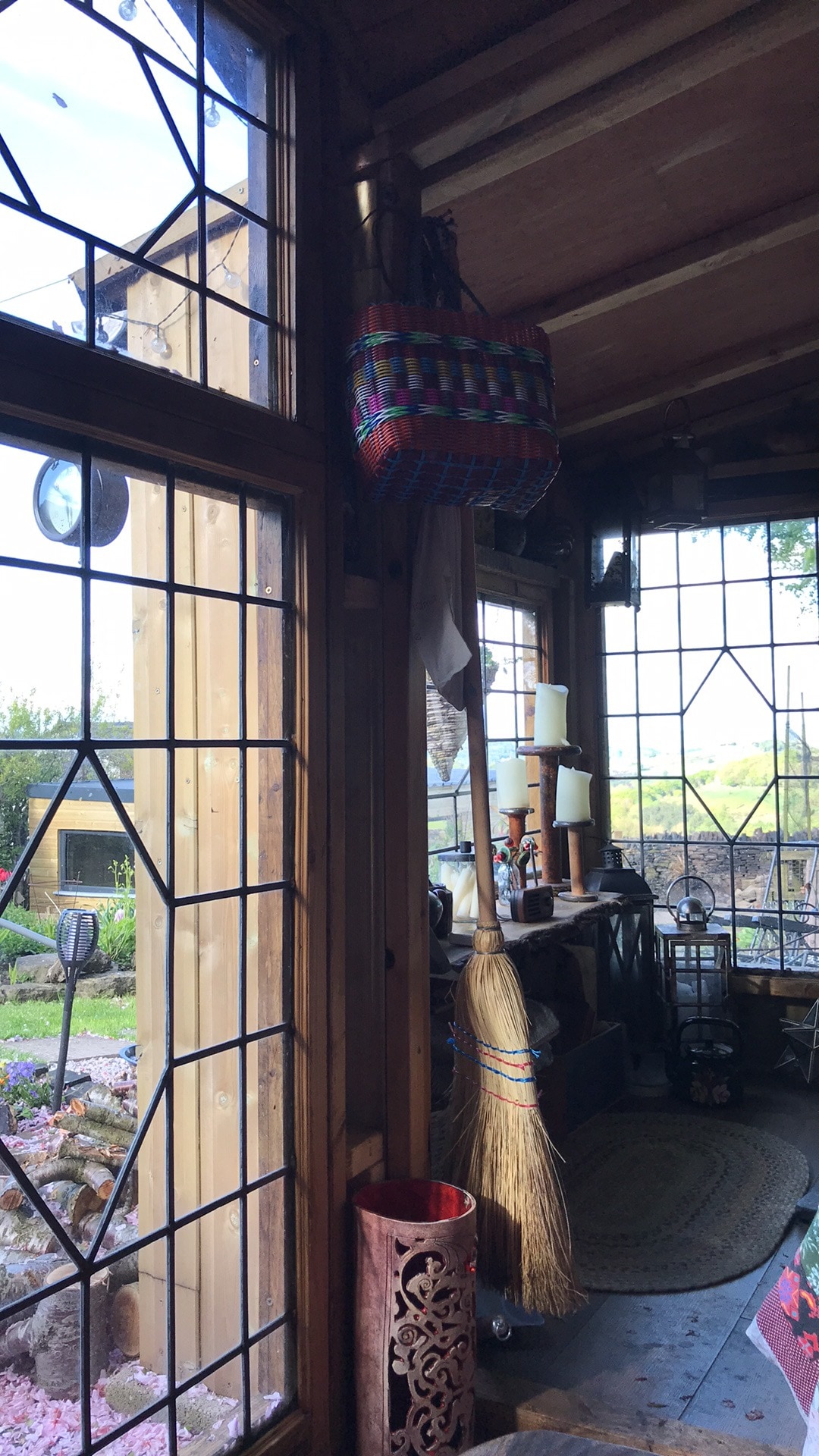 The interior features large windows, allowing natural light to fill the space. A colorful woven basket hangs from the ceiling, while wooden beams and decorative items create a cozy atmosphere. A broom and a decorative piece are visible near the floor, enhancing the rustic charm.