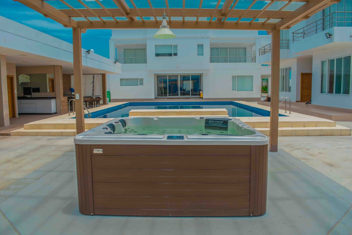 A hot tub is positioned under a wooden pergola, providing an inviting space for relaxation. The backdrop features a modern house with large windows and a nearby swimming pool, creating a harmonious outdoor area. The hardscape is well-maintained, enhancing the overall appearance.